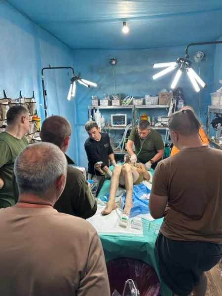 Чоловік, одягнений лише в шорти, лежить на ліжку, оточений шістьма іншими чоловіками. На стінах стоять полиці з різними медичними предметами, а також медичні предмети лежать на ліжку поруч із чоловіком.