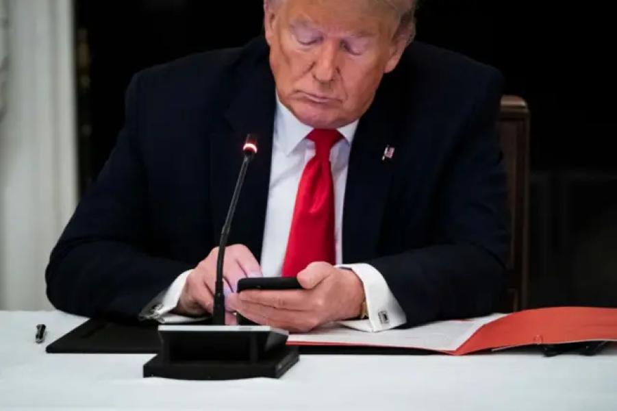 Дональд Трамп сидить за столом у, ймовірно, конференц-залі. Перед ним стоїть мікрофон, на столі лежить аркуш паперу в папці. Сам Трамп дивиться в екран смартфона, який тримає у руках