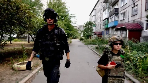 Ліворуч крокує високий чоловік у чорній формі, шоломі з навушниками та темних окулярах, із рацією та спорядженням на жилеті. Праворуч – молодша на вигляд дівчина в камуфляжному бронежилеті, шоломі та окулярах, у руках тримає червоний предмет (схоже на телефон або рацію). Вони йдуть вузькою асфальтованою дорогою між старими багатоповерхівками з балконами та зарослою зеленню обабіч