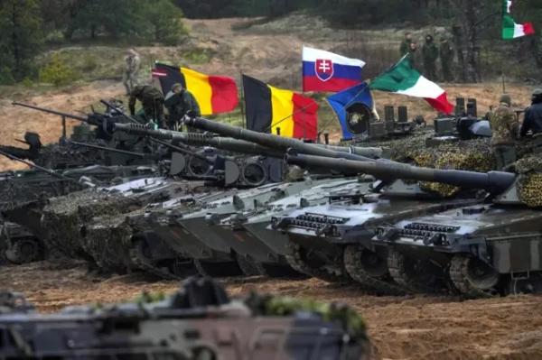 Ряд сучасних бойових танків стоїть на полі під час військових навчань. На баштах закріплені національні прапори Бельгії, Словаччини, Італії та інших країн НАТО. Військові у камуфляжі працюють біля техніки, на задньому плані видніються солдати серед дерев.
