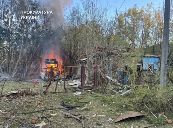 Ущент знищене сільське подвірʼя. Під деревами догорає автівка, від воріт залишився лише металевий каркас – і той погнувся, знищена стіна та дах невеликого будинку, всюди розкидані уламки матеріалів.