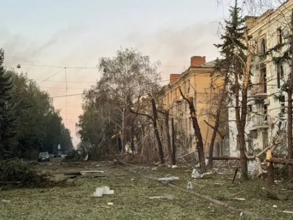 Вулиця повністю всипана листям і гілками дерев, що посікли уламки авіабомб. Від декількох дерев вздовж дороги залишилися тільки стовбури та пні, кілька – повалені на землю. На трьох поверхах багатоквартирного будинку вибиті всі вікна, покосилися рами. На дальньому плані припарковані легкові автомобілі