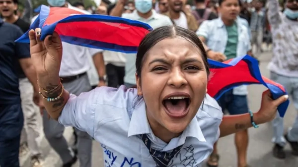 A young woman dressed in what appears to be a light blue school shirt, covered in graffiti, with a school tie tied loosely around her neck, shouts with her mouth wide open. She is carrying the Nepalese flag behind her head while a number of people walk down the street in the background, some wearing medical masks over their mouths. This protest takes place outside the Nepalese parliament in Kathmandu on 8 September 2025.