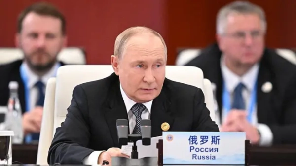 Володимир Путін, одягнений у темний костюм і чорно-білу краватку, сидить за столом на саміті і говорить у мікрофон. На табличці перед ним на столі написано слово &quot;Росія&quot; китайською, російською та англійською мовами.