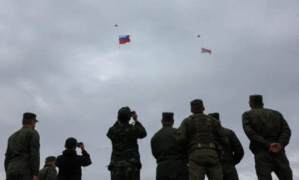 Квадрокоптери здійняли в небо російський і білоруський прапори. На передньому плані восьмеро військових, спиною до камери, дивляться у небо та знімають на телефони