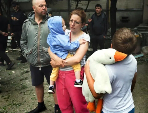Жінка у рожевих штанях триває на руках дитину у синій куртці. Хлопчик років 9-10 тримає м&#x27;яку іграшку-гуся і стоїть спиною до камери. Чоловік у зеленій куртці дивиться у бік. 