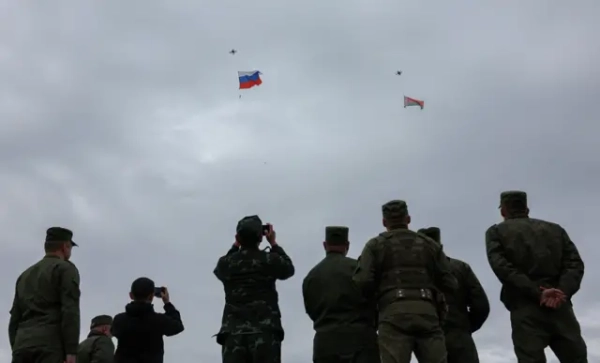 Квадрокоптери здійняли в небо російський і білоруський прапори. На передньому плані восьмеро військових, спиною до камери, дивляться у небо та знімають на телефони