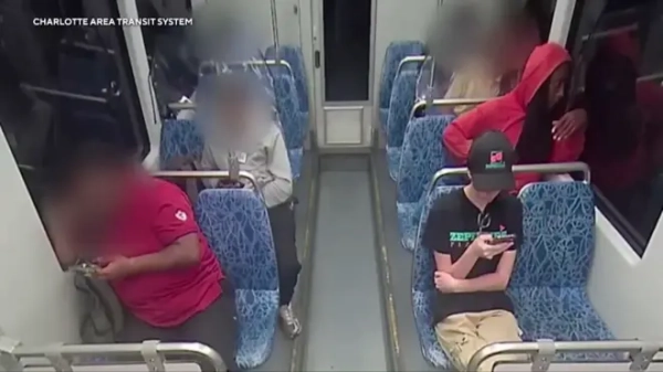 Камера відеоспостереження показує жінку в чорній бейсболці, футболці та штанах, яка приховує обличчя та дивиться телефоном у поїзді, а позаду неї сидить чоловік у червоній толстовці з капюшоном. 