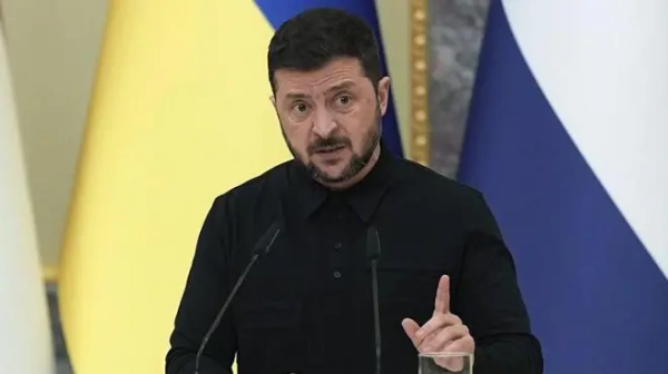 Володимир Зеленський у чорному костюмі стоїть за стійкою на фоні прапорів 