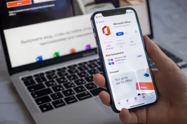 Людина тримає смартфон з Microsoft Office на екрані та ноутбук на столі на задньому плані