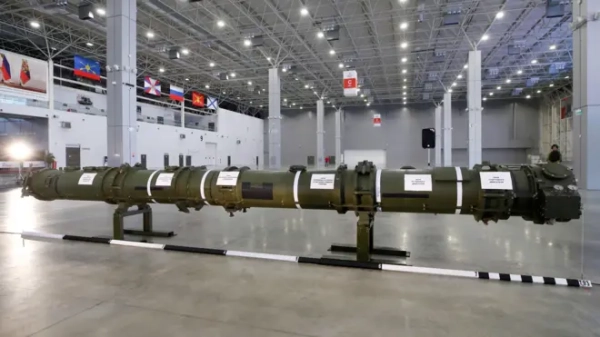 У великому виставковому павільйоні стоїть довга зелена ракета, розібрана на секції та встановлена на підставках. На корпусі видно білі смуги та таблички з написами. Позаду – порожній простір із високою стелею, прапорами різних країн і кількома людьми вдалині.