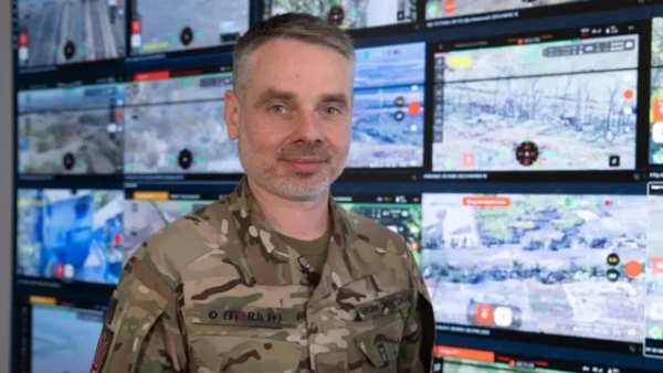 Юрій Мироненко стоїть у військовій формі перед стіною екранів, на яких зображені поля, чагарники та кілька будинків. Усі сцени з російсько-українського фронту