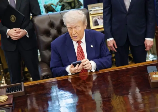 Трамп за столом Овального офісу тримає двома руками смартфон, сидячи у шкіряному кріслі. По обидва боки від нього – чоловіки в костюмах, їхні обличчя поза кадром