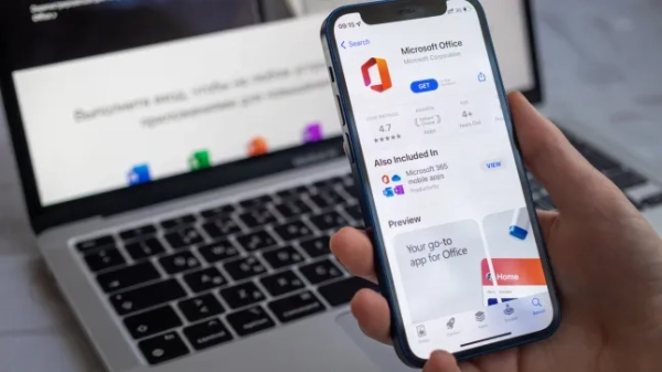 Людина тримає смартфон з Microsoft Office на екрані та ноутбук на столі на задньому плані