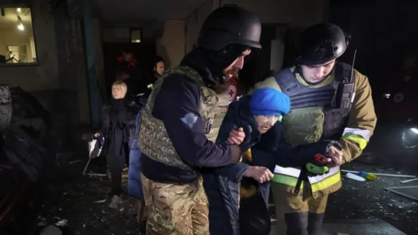 Двоє українських рятувальників підтримують літню жінку під час евакуації з місця російського удару по багатоповерховому житловому будинку в Харкові, північно-східна Україна, 19 листопада 2025 року. Жінка виглядає засмученою і нахилилася вперед. На ній синя шапка та зимова куртка.