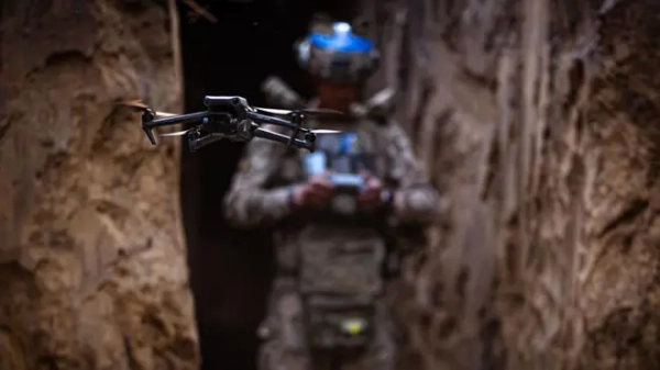 Розвідувальний дрон у повітрі між стінами окопу, на тлі в розмитті його оператор у військовій формі та екіпіруванні з синіми стрічками