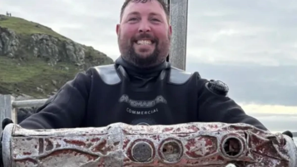 A man in a black wetsuit smiles as he holds a cylindrical metal object believed to be a sonobuoy. He is stood on a boat with, the sea and a portion of land can be seen behind him. He has short brown hair and a brown goatee style beard.