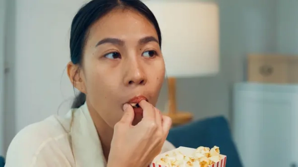 Close up crop of a stock photo showing a person&#x27;s face as they eat popcorn with their hands at home in the living room.