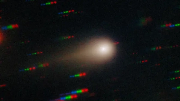 Комета 3I/Atlas зображена як яскравий центр, оточений туманною кулею із хвостом ліворуч; на задньому плані видно смуги синіх, зелених та червоних точок, що представляють зірки, що рухаються відносно комети