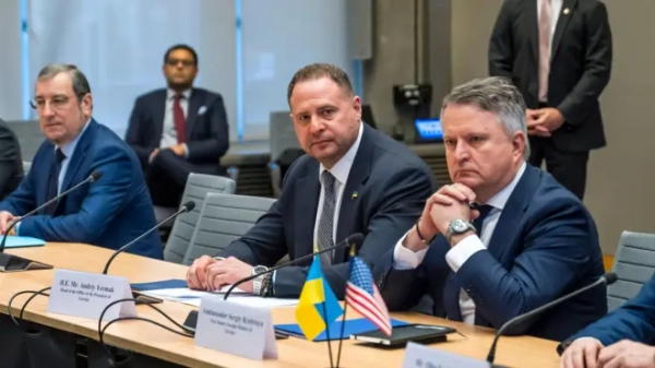 На фото кілька чоловіків у ділових костюмах сидять за довгим переговорним столом. На передньому плані Єрмак – який дивиться майже в камеру, та Кислиця – який сидить із складеними перед обличчям руками, зосереджений. На столі перед ними стоять маленькі прапорці України та США. Позаду видно інших учасників зустрічі та охоронця, який стоїть біля стіни.