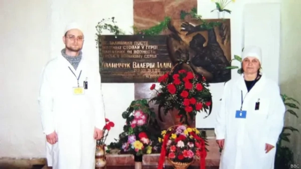 Чоловік і жінка у білих халатах та шапочках стоять по обидва боки меморіальної плити, уквітчаної великими букетами червоних, рожевих і жовтих квітів. На плиті – рельєфна скульптурна композиція та напис українською мовою: &quot;Не залишили пости, мужньо стояли у герці, памʼятник їм вознести треба у кожному серці. Ходимчук Валерій Ілліч&quot;