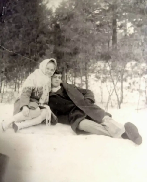 На чорно-білому знімку молоде подружжя сидить разом на снігу серед зимового лісу. Жінка в хустці та теплому пальті сидить збоку й ніжно схиляється до чоловіка. Чоловік у темному пальті напівлежить поруч, водночас трохи обіймаючи її. Обоє виглядають спокійними та близькими, позують на фоні високих засніжених дерев