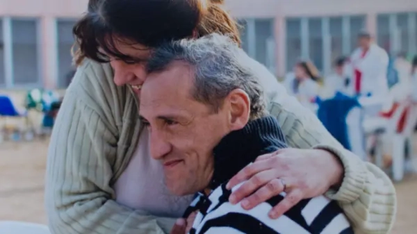 Жінка обіймає за плечі чоловіка, який сидить боком до камери, вони обоє посміхаються