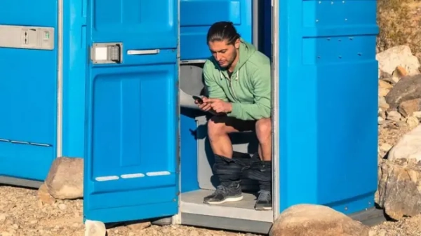 A man sitting on the toilet in a portable toilet in the middle of the Death Valley desert, in the United States. He has his trousers rolled around his ankles and is reading his phone.  The toilet door is wedged open by a rock and another portable toilet is next to hium, with the door closed. Rocks are scattered around and there are barren hills in the background.