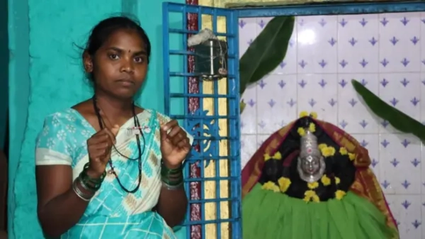 Шилпа тримає намисто, яке носять девадасі. Вона дивиться в камеру з нейтральним виразом обличчя, одягнена в бірюзове сарі та стоїть поруч зі святилищем індуїстського бога.