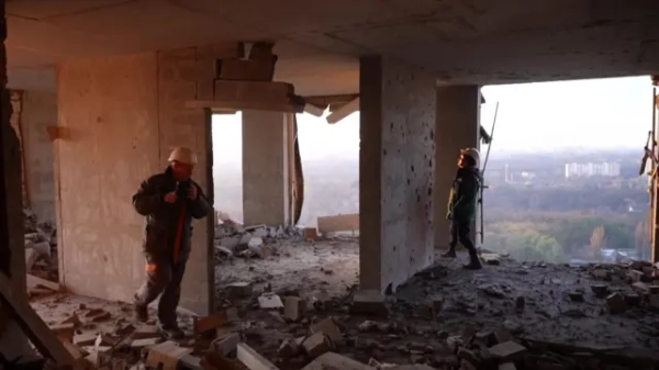 Двоє пожежників у захисному спорядженні проходять крізь зруйновану будівлю високо над землею. Вікна вибиті, крізь щілини в стіні видно будівлі та дерева.