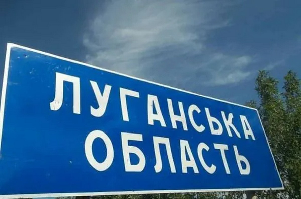 Дорожній знак на фоні зелених дерев та неба: напис "Луганська область" білими літерами на синьому фоні.