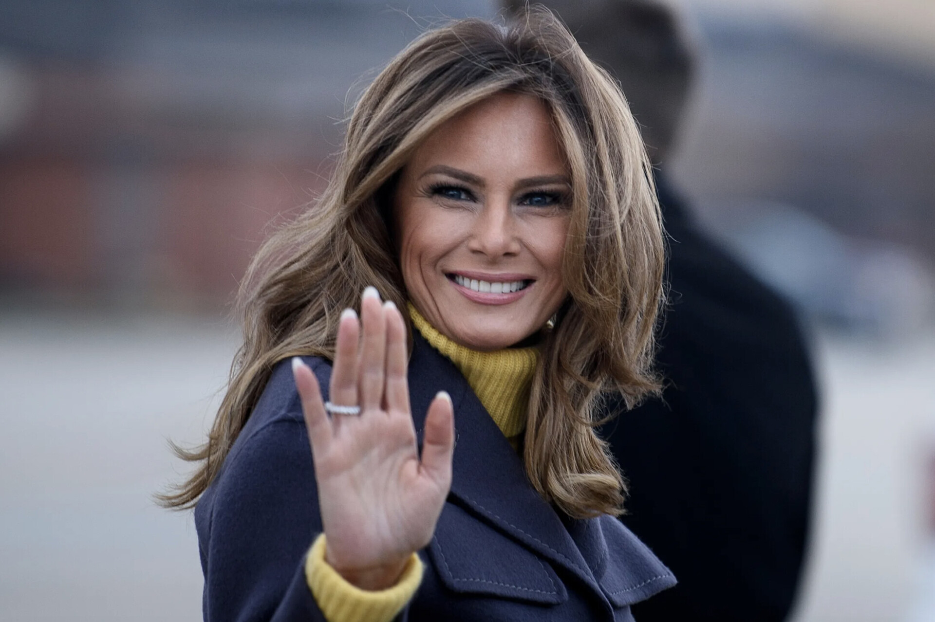 Меланія Трамп усміхається та махає рукою на публічному заході. Вона одягнена в темне пальто й гірчичний светр.