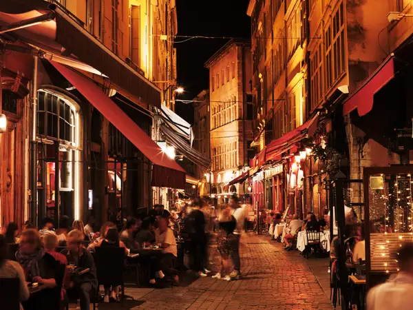 Warmly lit narrow cobblestone street with outdoor restaurant seating and blurred diners at night.