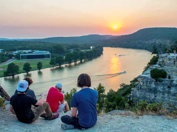 Група друзів сидить на краю скелі з річками та пагорбами на тлі заходу сонця.