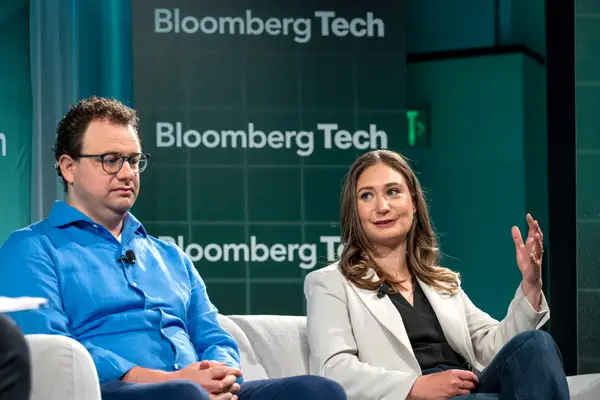 Двоє спікерів сидять на сцені на тлі зеленого фону BloombergTech під час технологічної події.